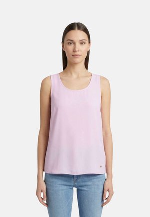 Donna con capelli lisci e scuri indossa un top rosa chiaro senza maniche e jeans blu, in piedi su uno sfondo bianco semplice.
