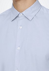 Camicia azzurra chiara con colletto classico, texture liscia e bottoni bianchi in plastica, con cuciture ordinate e senza fantasie.