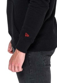 Felpa nera in tessuto testurizzato, con accento del logo rosso sul polsino sinistro. La ripresa ravvicinata mostra la manica, la mano e jeans scuri.