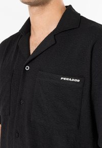 Man draagt een gestructureerd zwart overhemd met knopen, een kraag en een borstzak met de tekst "PEGADOR" geborduurd in witte letters.