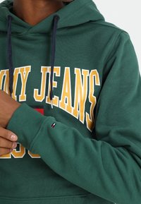 Green hoodie with “TOMMY JEANS” in yellow text, featuring a front pocket, drawstrings, ribbed cuffs, and a small logo on the sleeve.