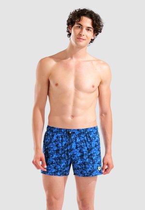 Junger Mann mit lockigem Haar, der blaue gemusterte Badeshorts trägt, steht vor einem einfarbigen hellgrauen Hintergrund.