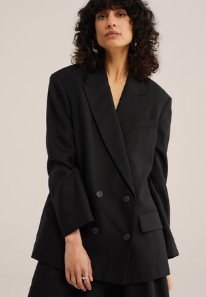 Donna con capelli ricci scuri che indossa un blazer nero doppiopetto oversize su sfondo beige, con orecchini a cerchio d'argento e anelli.