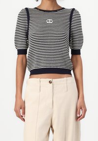 Svetr pletený v námořnické modré a smetanové pruhy, s krátkými puff rukávy, žebrovanými manžetami a stříbrným logem na hrudi. Stylizováno s béžovými kalhotami.