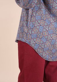 Chemise à manches longues à motifs avec des designs géométriques bleus, oranges et verts, associée à un pantalon rouge foncé uni sur un fond beige.