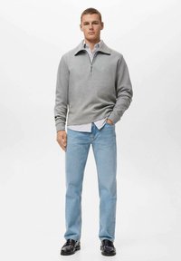 Grijze zip-nek sweatshirt over een lichtblauw gestreept shirt, gecombineerd met lichtblauwe jeans en zwarte leren schoenen, staande tegen een witte achtergrond.