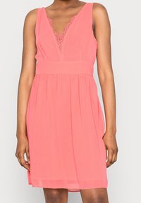 Robe sans manches corail avec un décolleté en V, des détails en dentelle à l'avant et une taille cintrée menant à une jupe évasée. Tissu doux et léger.