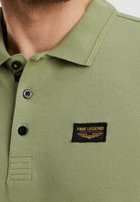 Olivengrünes Poloshirt aus strukturiertem Stoff, mit einem schwarzen Etikett mit goldener Schrift und Logo. Die Knöpfe sind schwarz und rund.