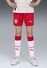Röd fotbollskit med en vit tröja som har subtila mönster, röda shorts med en logotyp, röda och vita strumpor samt grå fotbollsskor.
