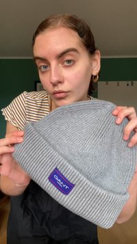 Grå strikket hue med ribbet tekstur og foldet skygge, der har et lilla og blåt GANT logo patch.