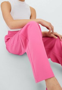 Person som bär en ärmlös vit topp och ljusrosa vida byxor, sittande med händerna vilande på det uppdragna knäet mot en ljus bakgrund.