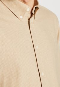 Chemise beige à boutons avec un motif quadrillé subtil portée par une personne, avec un col boutonné et des boutons blancs visibles à l'avant.