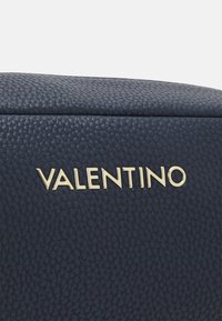 Lähikuva laivansinisestä nupukkinahasta, jossa kullanvärinen Valentino-logoteksti on kohokuvioitu pinnalle.
