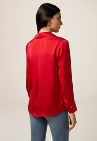 Blouse en satin rouge avec des manches longues, un col de chemise et des boutons aux poignets. Texture lisse, coupe ajustée et détails au dos.