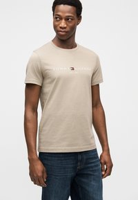 Μπεζ βαμβακερό μπλουζάκι με κοντά μανίκια, με λευκά γράμματα "TOMMY HILFIGER" και λογότυπο σε κόκκινο, λευκό και μπλε. Συνδυάζεται με σκούρο τζιν παντελόνι.