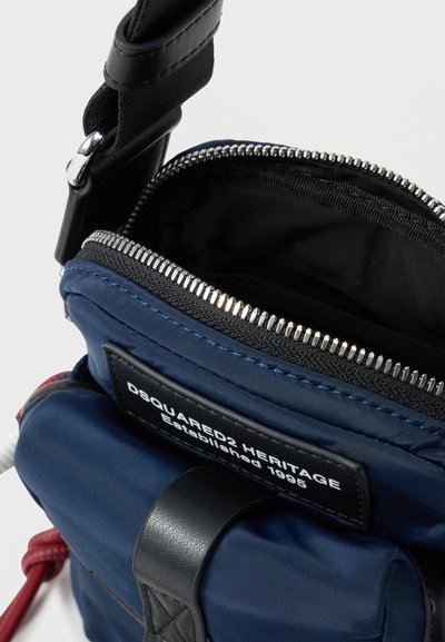 Μπλε υφασμάτινη τσάντα χιαστί με μαύρο λουρί, ανοιχτό φερμουάρ και ετικέτα "DSQUARED2 HERITAGE Established 1995" στην μπροστινή τσέπη.