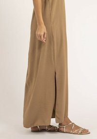 Robe maxi beige avec une fente sur le côté, fabriquée en tissu léger et texturé. Associée à des sandales plates avec des lanières métalliques.