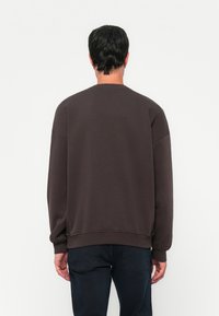 Man wearing a dark brown long-sleeve sweatshirt and dark jeans, standing facing away against a plain white background.