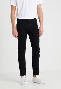 Man som bär slim-fit svarta jeans, vita sneakers och en vit kortärmad t-shirt, som står mot en enfärgad ljus bakgrund.