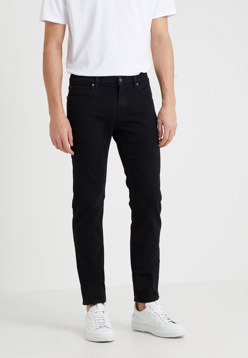 Man som bär slim-fit svarta jeans, vita sneakers och en vit kortärmad t-shirt, som står mot en enfärgad ljus bakgrund.