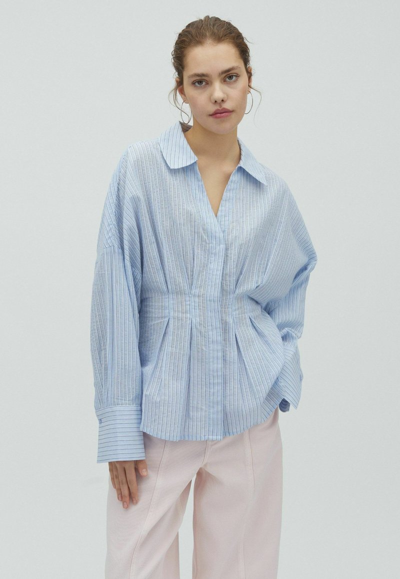 Femme aux cheveux attachés porte une blouse oversize bleu clair à fines rayures et un pantalon rose clair, debout devant un fond uni.