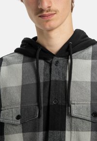 Camisa de cuadros con cuadrados grises y negros, con capucha negra, cierre frontal de botones y dos bolsillos en el pecho. Tela suave y texturizada.