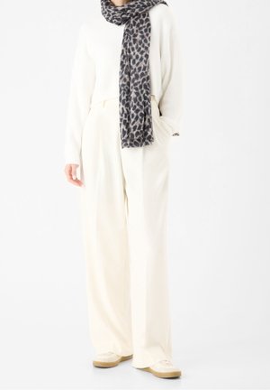 Personne portant un pantalon blanc à jambes larges, un pull blanc, une écharpe à imprimé léopard et des baskets blanches, debout les mains dans les poches.