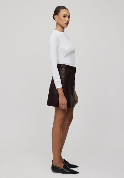 Maglietta bianca a maniche lunghe abbinata a una gonna in pelle marrone scuro. La gonna presenta tasche e una texture liscia, completata da scarpe nere senza lacci.