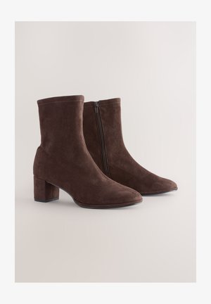 Cizme maro din piele întoarsă cu toc gros, prevăzute cu fermoar lateral și design cu vârf ascuțit. Textură netedă cu detalii subtile de cusături.