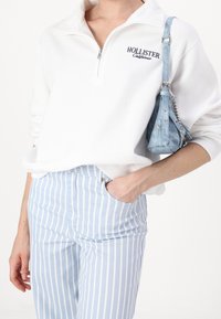 Vitt fleece tröja med halvhög krage och "Hollister California"-logotyp. Ljust blå och vitrandig byxa med fickor. Liten blå handväska.