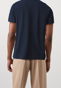 Camiseta de algodón azul marino con mangas cortas, cuello redondo y dobladillo recto, combinada con pantalones beige, vista desde la parte posterior.