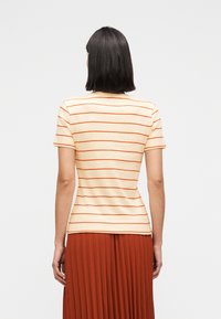 Γυναίκα με ίσια σκούρα μαλλιά που φοράει μια κρεμ κοντομάνικη μπλούζα με λεπτές πορτοκαλί οριζόντιες ρίγες και μια καφέ πλισέ φούστα.