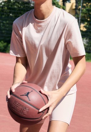 Osoba w jasnoróżowym stroju sportowym trzyma brązową piłkę do koszykówki na otwartym boisku z zielonym ogrodzeniem w tle.