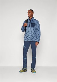 Zils rakstains fliisa pulovers ar pusgaru rāvējslēdzēja apkakli, kombinēts ar tumšziliem biksēm ar gaiši ziliem akcentiem un vieglām pārgājienu kurpēm.