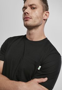 Fekete, rövidujjú póló, pamutból készült, elülső zsebbel és egy kis zöld címkével az ujjon. Zökkenőmentes textúra.