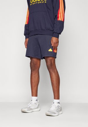 Personne portant un short de sport Adidas bleu marine et un hoodie assorti avec des bandes rouges et jaunes, des baskets blanches Adidas et des chaussettes.