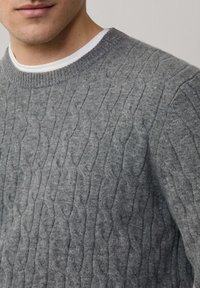 Maglione grigio lavorato a maglia con motivo a treccia, scollatura rotonda e polsini a coste. Realizzato in un materiale morbido e testurizzato, adatto per essere indossato a strati.