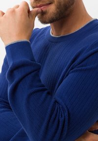 Blå ribbad tröja med texturerat tyg, rund hals och kontrasterande grå detaljer vid ärmsluten. Nära bild på ärmarna och halsringningen.