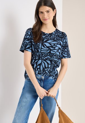 Mujer con blusa de manga corta azul con estampado y vaqueros, sosteniendo un bolso marrón con ambas manos frente a un fondo neutro.