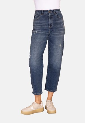 Person wearing high-waisted, slightly distressed blue jeans and beige sneakers with light brown soles, standing against a plain background.
