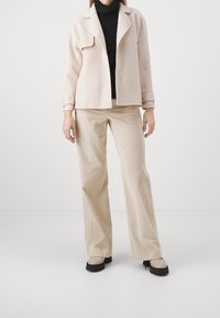 Vaaleanpunainen takki mustan poolopaidan päällä, yhdistetty beigen värisiin ribbineulottuihin leveälahkeisiin housuihin ja vaaleanharmaisiin kenkiin, joissa on mustat pohjat.