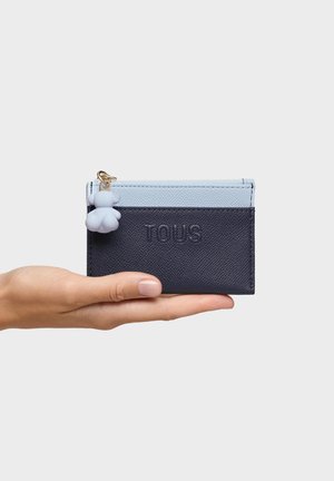 Dłoń trzyma granatowo-niebieski etui na karty z wytłoczonym logo "TOUS" oraz małym białym breloczkiem w kształcie misia przypiętym do zamka.
