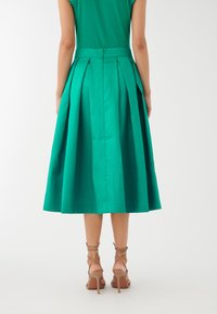 Zelena plisirana midi krilo iz gladkega materiala, ki ima prilegajoč pas in razširjen silhuet. Nošeno z bež čevlji na peto z trakovi.