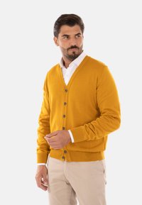 Cardigan giallo senape da uomo realizzato in maglia, con scollatura a V, chiusura con bottoni e polsini a costine. Indossato sopra una camicia bianca.