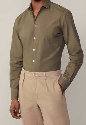 Uomo che indossa una camicia verde oliva con bottoni e pantaloni beige plissettati con mani rilassate, sfondo beige.