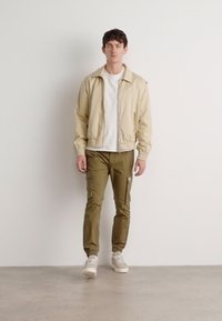 Lichtbeige zip-jack over een wit shirt, gecombineerd met olijfgroene cargobroek en lichte sneakers, staande tegen een effen achtergrond.
