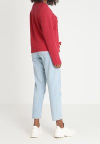 Punainen ribbineulottu neule, jossa on teksturoitu etuosa ja vyötäröllä solmittava yksityiskohta, yhdistetty vaaleansinisiin farkkuihin ja valkoisiin lenkkareihin.