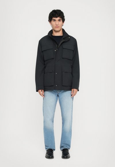Giacca nera isolata con quattro tasche frontali, chiusura a zip e colletto alto. Indossata con jeans azzurri chiari e calzature nere.