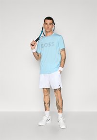 Chemise bleu clair à manches courtes avec "BOSS" en gris, assortie à un short blanc. Le modèle tient une raquette de tennis, portant des baskets blanches et des bandes de poignet.