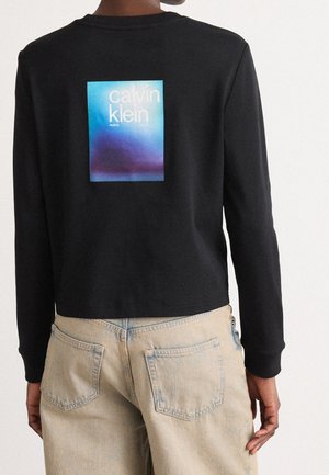 Osoba nosząca czarną bluzkę z długim rękawem z niebiesko-fioletowym logo Calvin Klein na plecach, w połączeniu z jasnobeżowymi dżinsami.